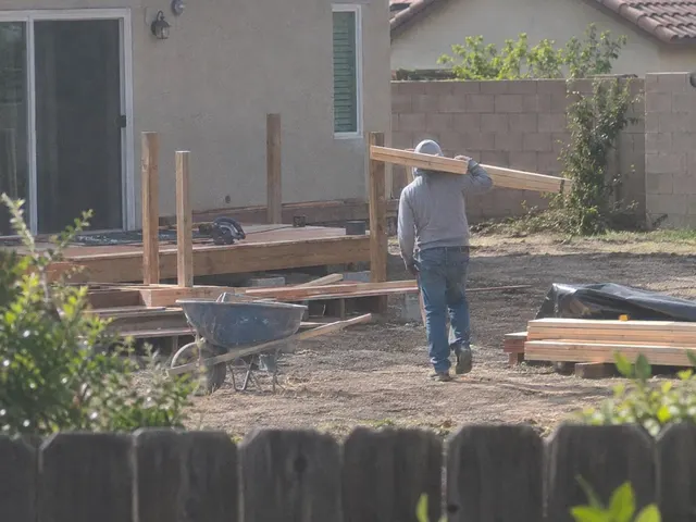 The same man carrying long planks across a building site