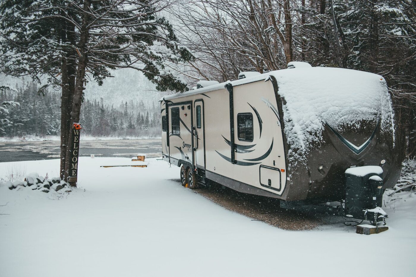 Travel trailer covered in snow parked beside a river in a winter forest